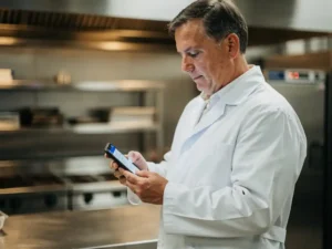 Voedselveiligheidsinspecteur in witte jas controleert digitale checklist op smartphone in professionele roestvrijstalen keuken.