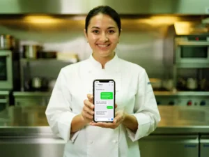 Voedingsveiligheidsexpert in witte koksjas toont WhatsApp-gesprek op smartphone in professionele roestvrijstalen keuken.